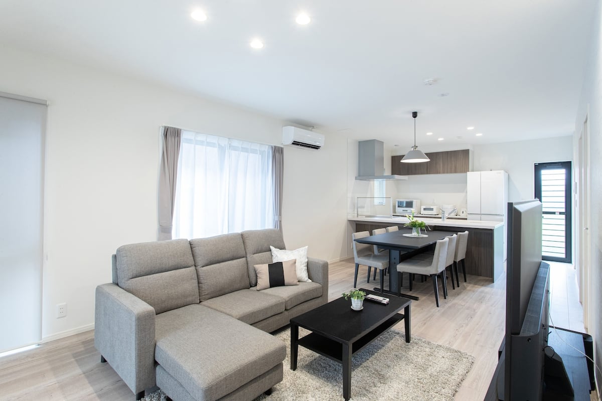 A modern living room with a sofa, coffee table, and TV. There is a dining area with a table and chairs. The kitchen is visible in the background with cabinets and appliances.