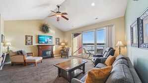 A spacious living room with a fireplace, comfortable seating, a flat-screen TV, and a large window offering a view of the outdoors. - Beachtop Condo at Delton Grand - Waterfront (Wisconsin Dells)