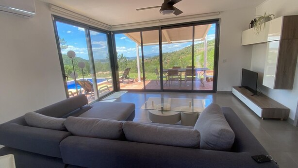 A modern living room with a large gray sofa, a coffee table, and a TV on a wooden stand. The room has large sliding glass doors leading to an outdoor patio with a pool and dining area. - Casa da Alfarrobeira-Villa 4+2P Pool & View (São Bartolomeu de Messines)