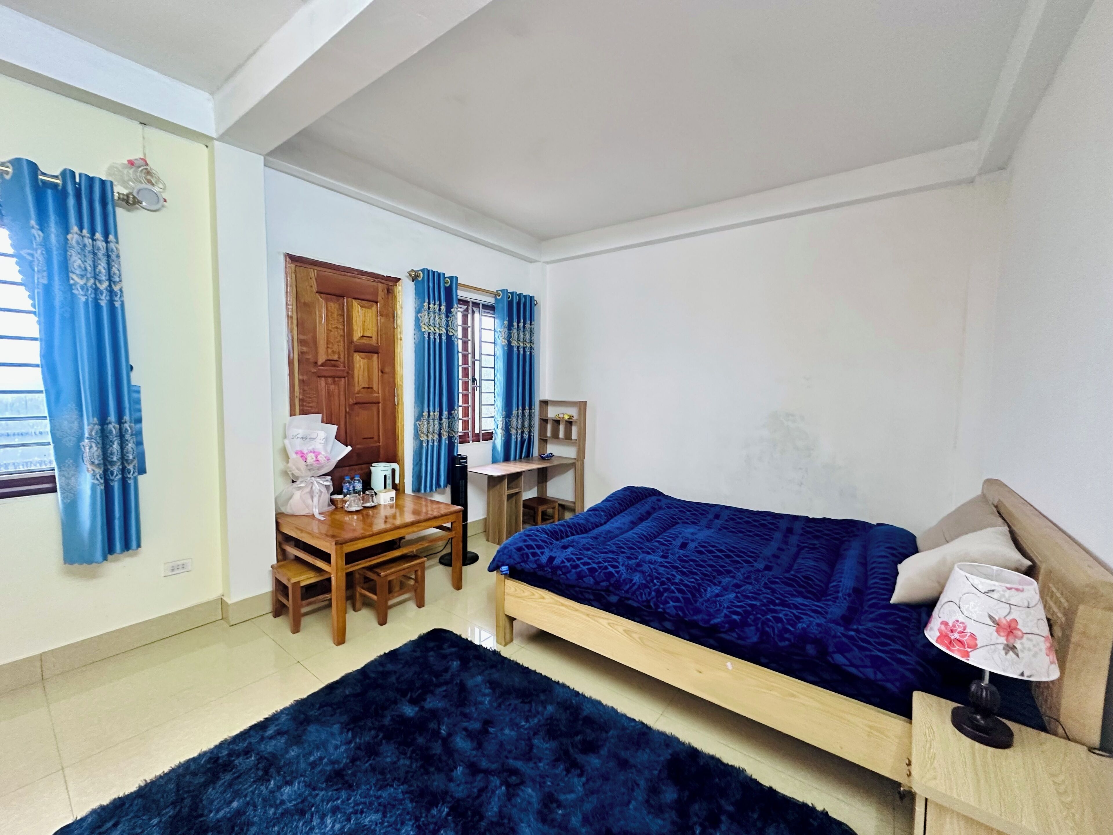 Un dormitorio con cama, escritorio y silla. Hay una ventana con cortinas, una puerta y una pequeña mesa con objetos encima.