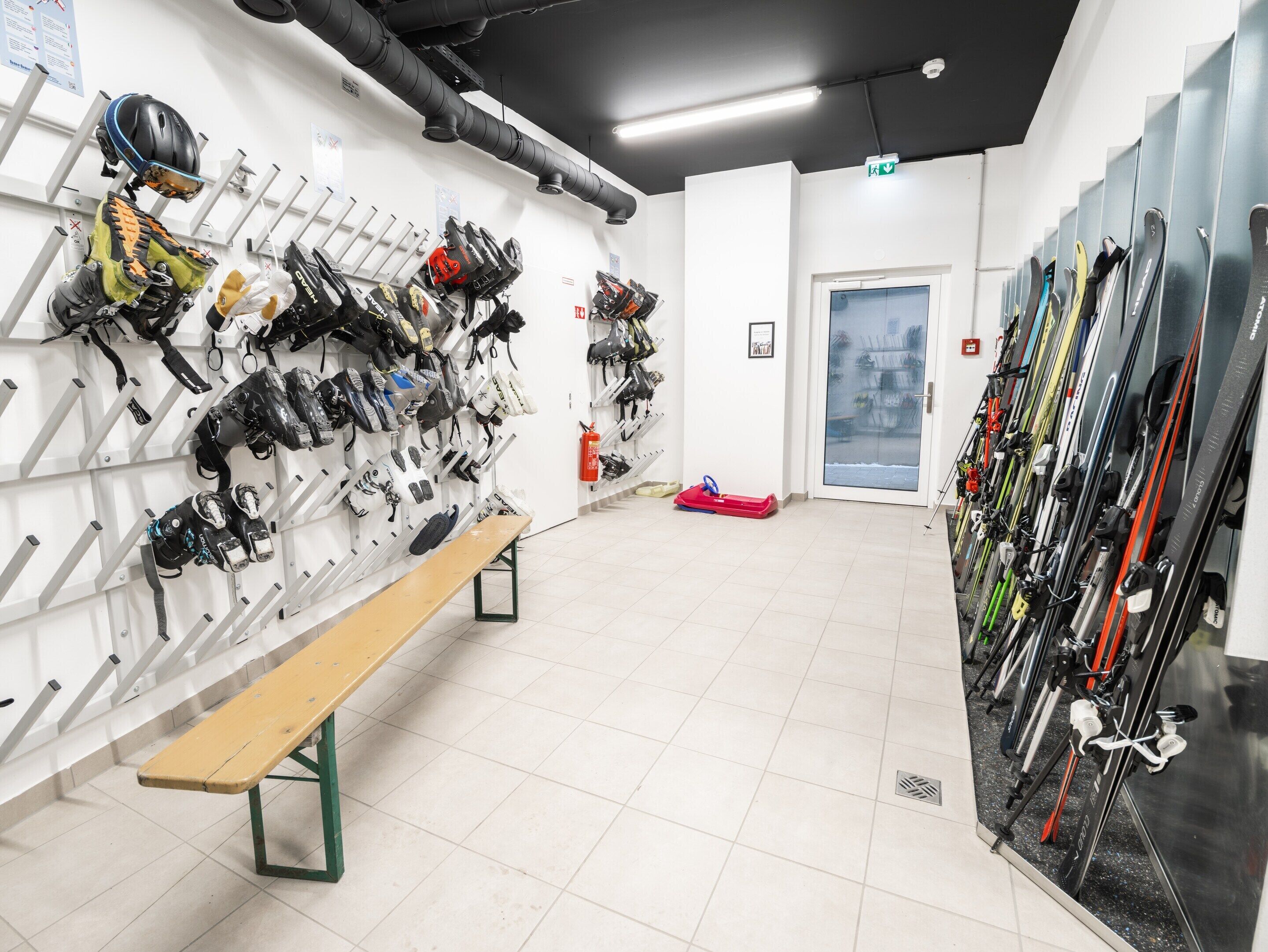 Een winkel voor ski- en snowboardverhuur, met rijen ski's en snowboards, een bankje en een brandblusser.