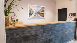 A reception desk with a wooden top and a textured grey panel. There is a framed landscape photo on the wall, a potted plant, and a vase with flowers. - Apartment in Fieberbrunn With Balcony (Fieberbrunn)