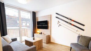A modern living room with a grey sofa, a wooden coffee table, and a TV mounted on a wooden wall. There are skis hanging on the wall and a balcony with a view of a building. - Apartment in Fieberbrunn With Balcony (Fieberbrunn)