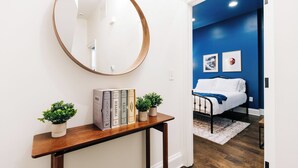A hallway with a wooden console table, a round mirror, and a bed in the adjacent room.
