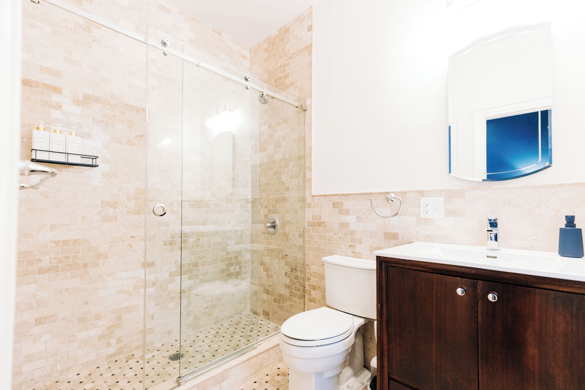 A bathroom with a glass shower enclosure, a white toilet, a wooden vanity with a mirror, and a white sink.