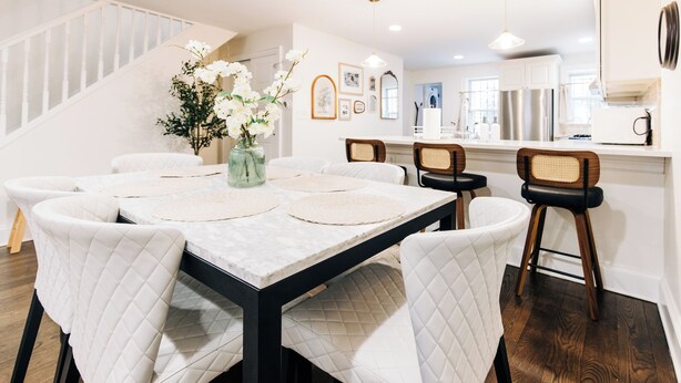 A dining area with a marble table, white quilted chairs, a vase with flowers, and a staircase in the background.