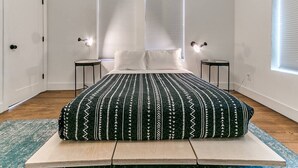 A bedroom with a bed, two bedside tables, a window with blinds, and a door.