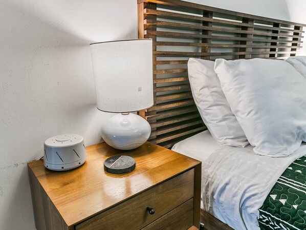 A neatly made bed with white bedding and pillows, a wooden nightstand with a lamp and a small device, and a wooden headboard with horizontal slats.