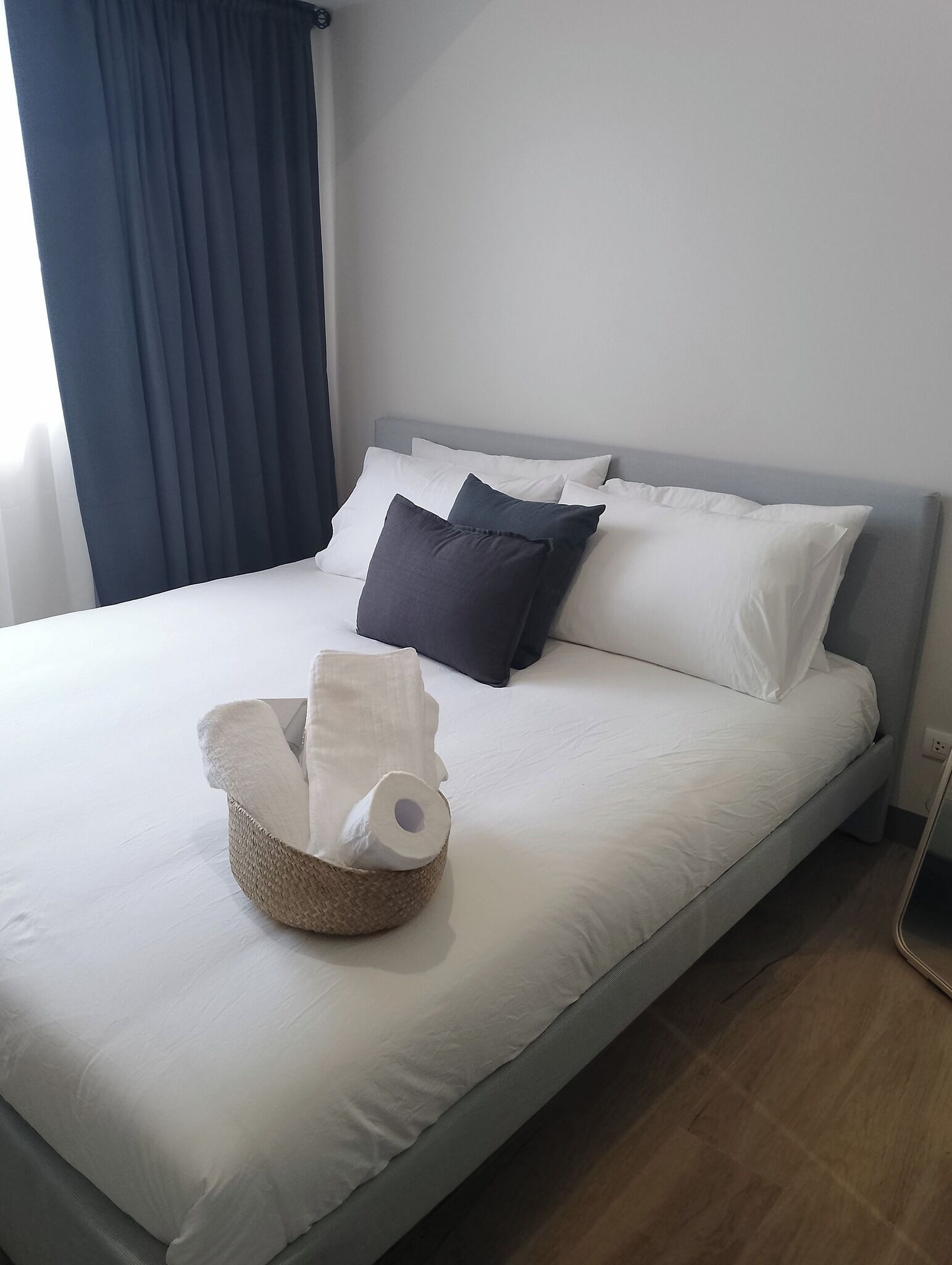Un lit bien fait, avec des oreillers blancs et gris foncé, un panier contenant un rouleau de papier toilette, et un rideau foncé partiellement tiré sur le côté.