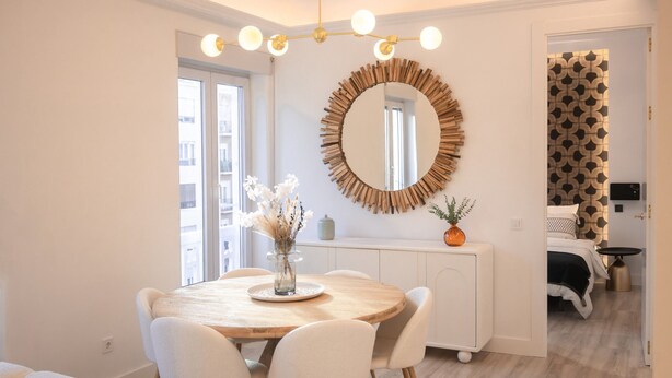 Un coin repas avec une table ronde en bois, des chaises blanches, un vase rempli de fleurs et un grand miroir accroché au mur.