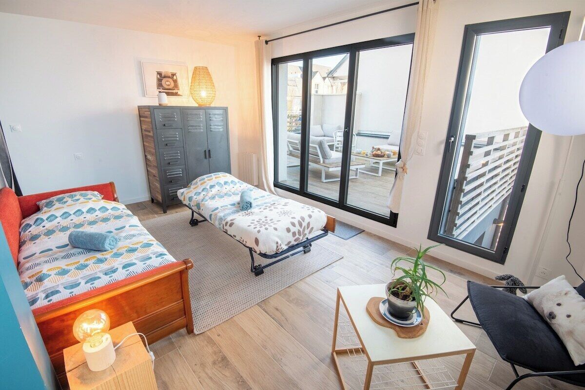 Habitación pequeña con cama, banca, una mesita con una planta y vistas al balcón exterior.