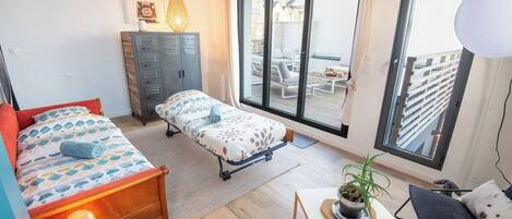Habitación pequeña con cama, banca, una mesita con una planta y vistas al balcón exterior.