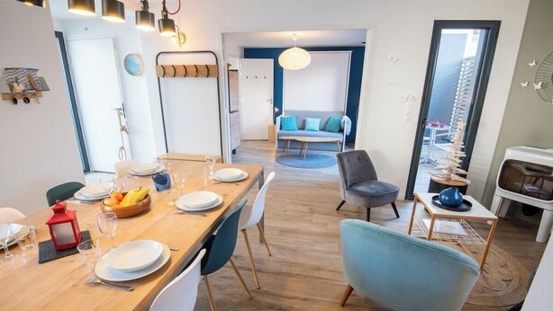 A modern dining area with a wooden table set for four, surrounded by white chairs. A blue armchair is positioned near the table, and a gray sofa is visible in the background. - Beautiful villa in Lomener-beach, restaurants, and shops within walking distance (Ploemeur)