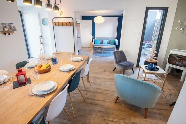 A modern dining area with a wooden table set for four, surrounded by white chairs. A blue armchair is positioned near the table, and a gray sofa is visible in the background. - Beautiful villa in Lomener-beach, restaurants, and shops within walking distance (Ploemeur)