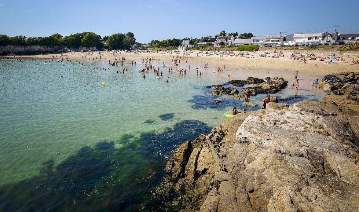 Une plage à l’eau claire, avec un rivage rocheux et des bâtiments en arrière-plan.