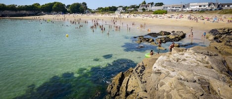 Una playa con agua cristalina, costa rocosa y edificios al fondo.