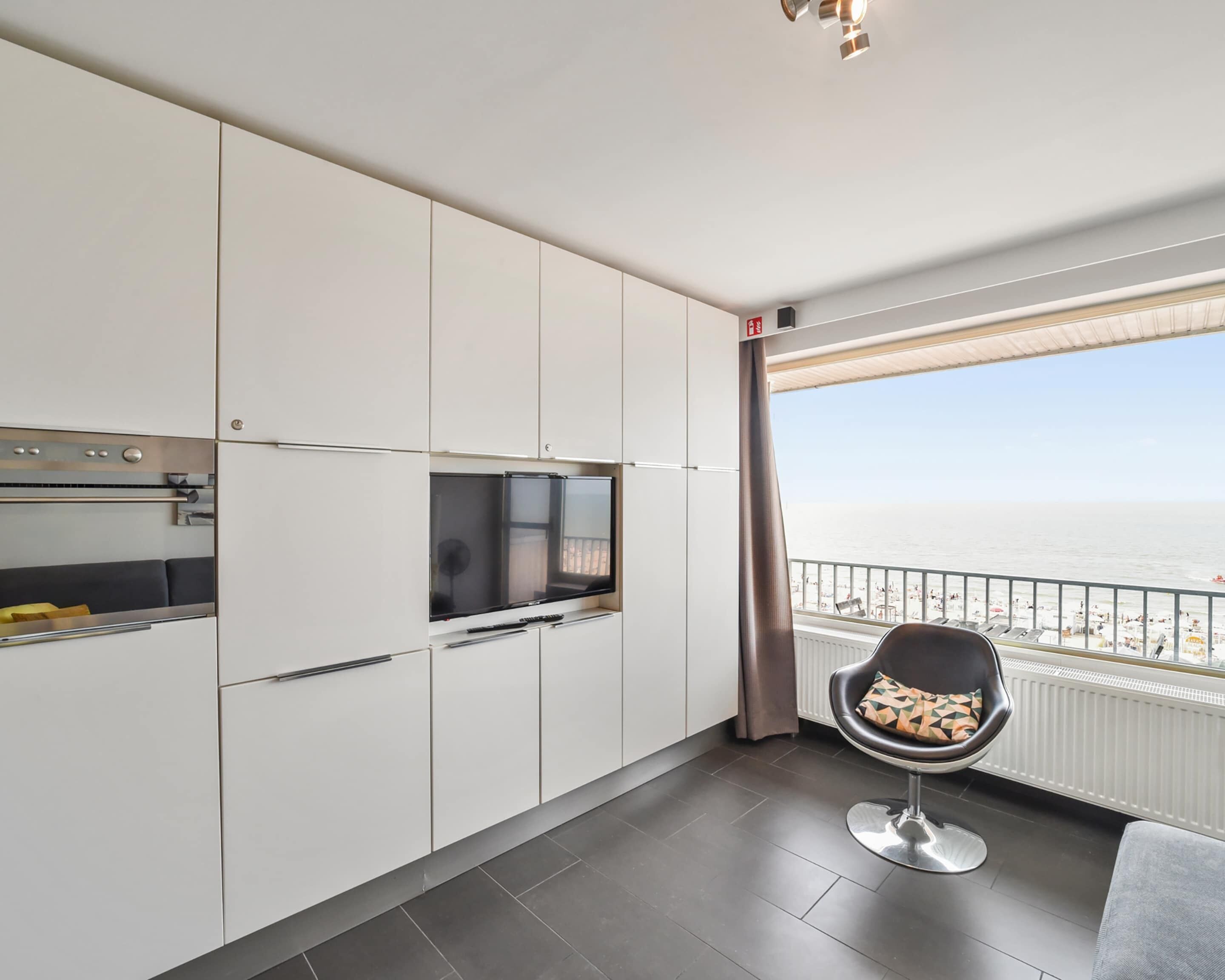 A modern kitchen with white cabinets, a built-in oven, and a large window overlooking a beach.