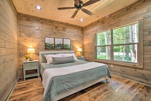 Un dormitorio con paneles de madera, una cama, dos mesitas de noche, un ventilador de techo y un ventanal con vistas a árboles.