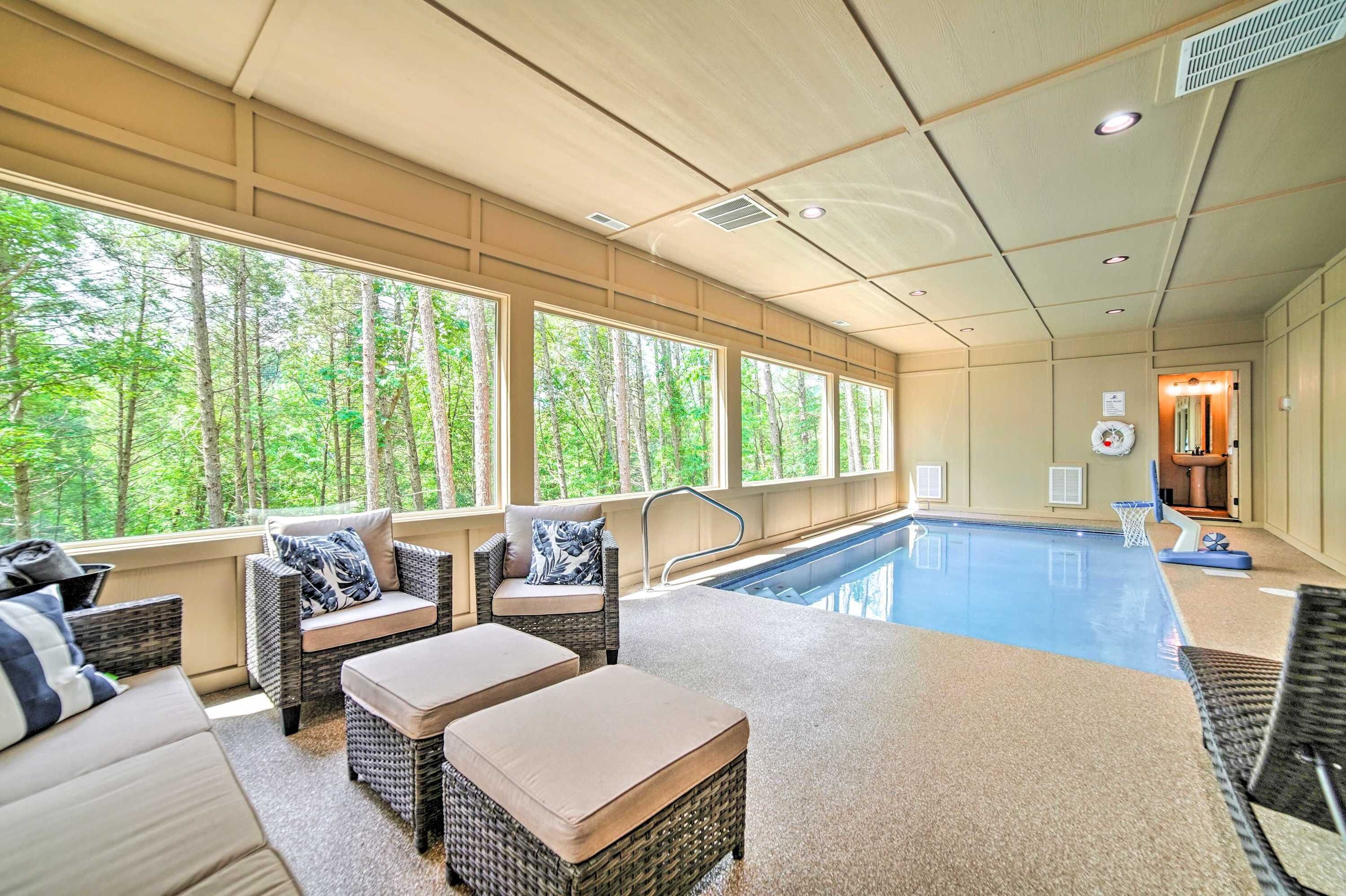 Zona de piscina interior con mobiliario de mimbre y amplias ventanas con vistas a un bosque.