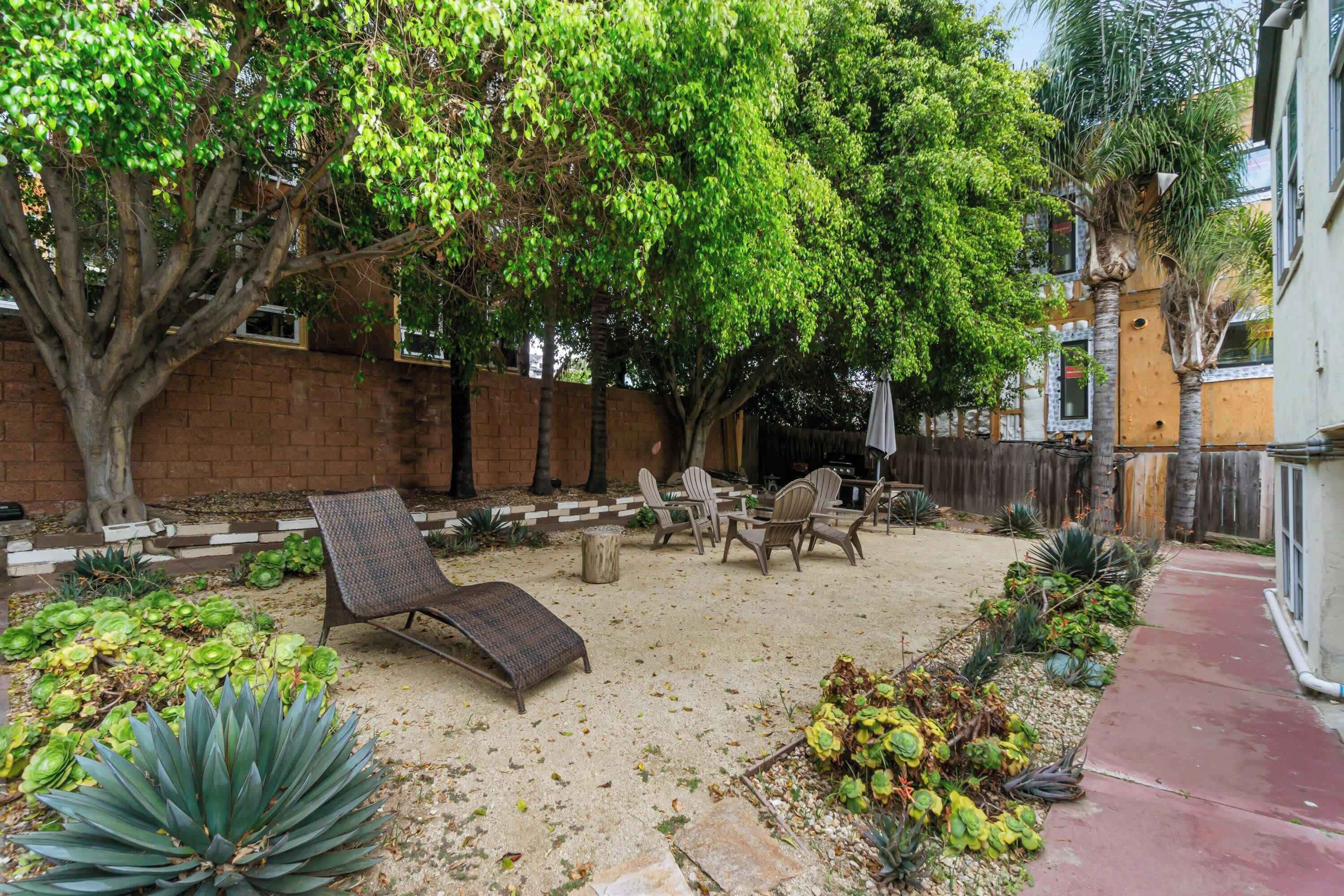 A backyard with a brick wall, a large tree, and a patio area with chairs and a lounge chair.