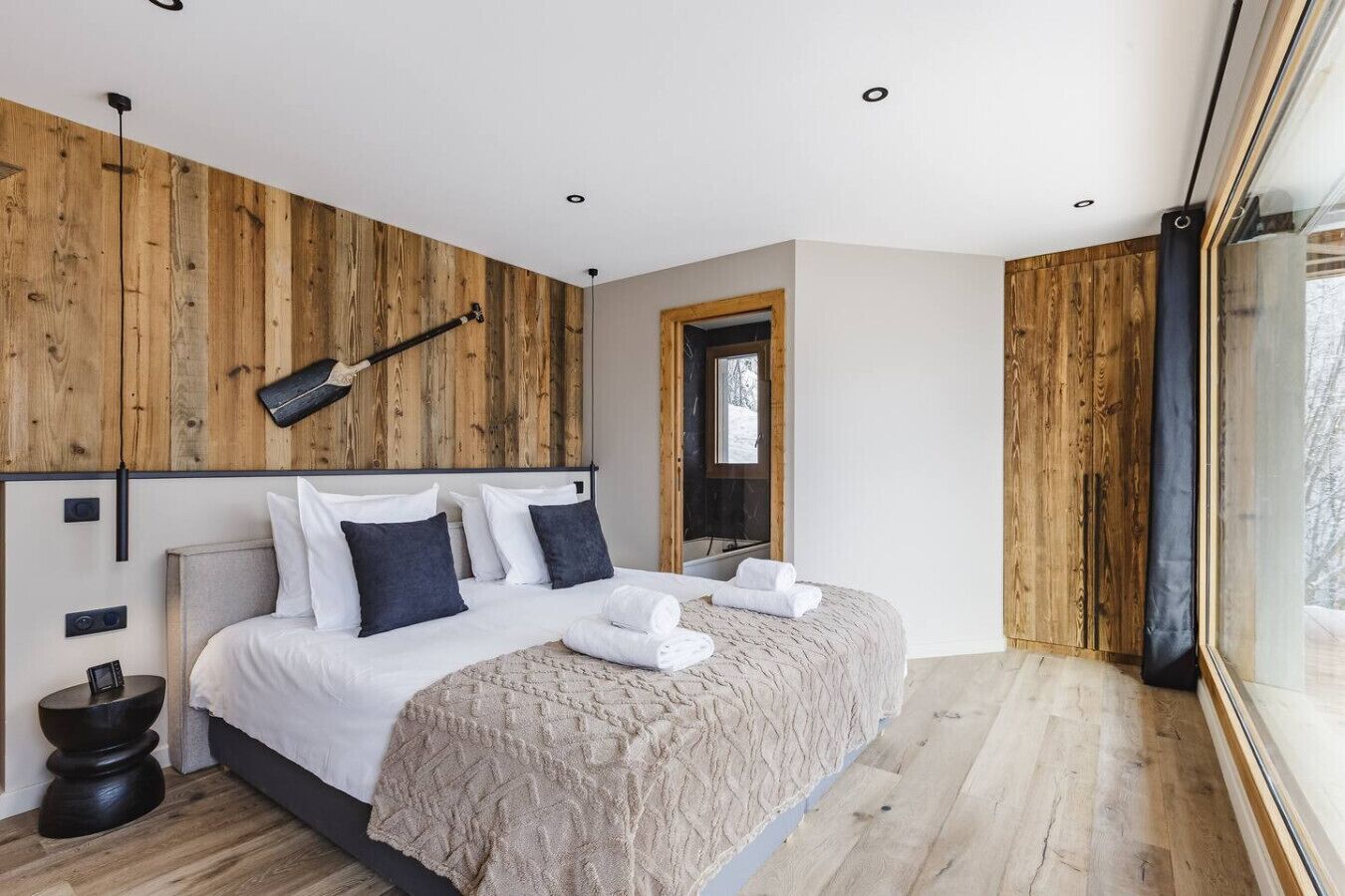 Una camera da letto moderna con una testiera in legno, un letto grande con biancheria da letto bianca, un comodino e una finestra con vista sugli alberi.