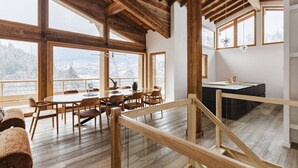 Un comedor amplio con techo alto y vigas de madera, ventanas grandes que ofrecen vistas del paisaje nevado, y una cocina moderna con encimera de madera y lámpara colgante.