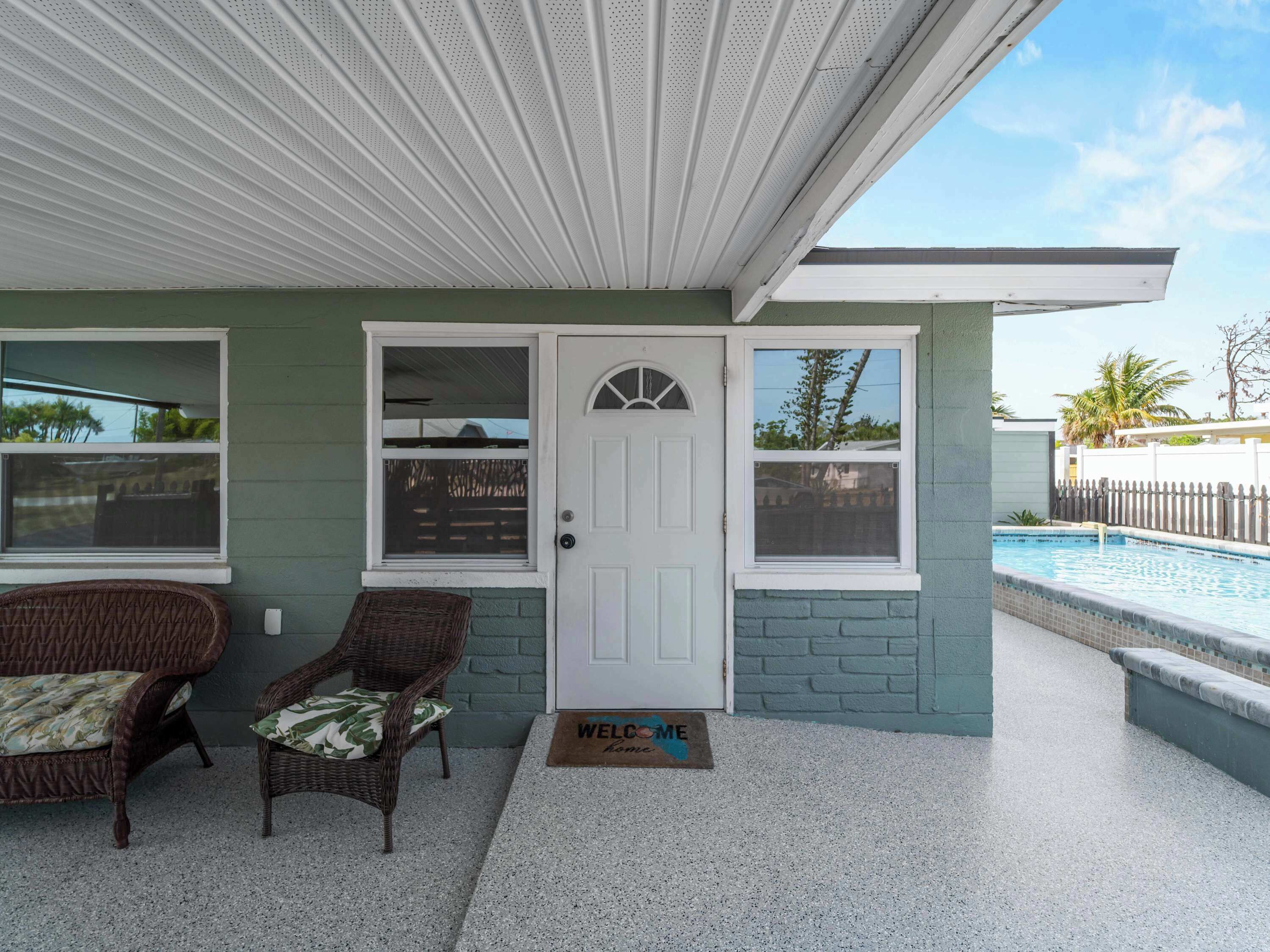 A house with a covered porch, two wicker chairs, a door with a welcome mat, and a swimming pool in the background.