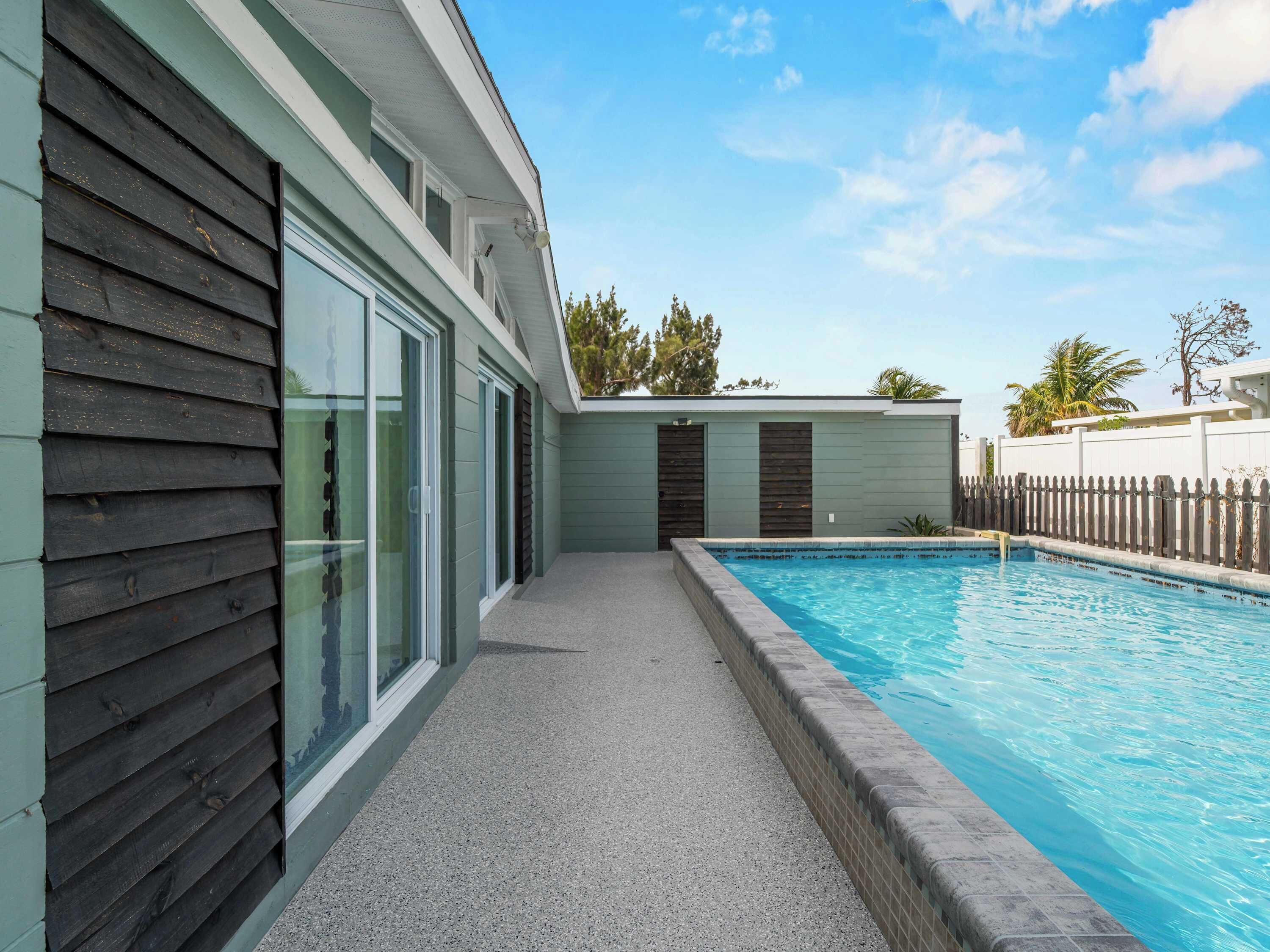 A modern outdoor swimming pool area with a concrete deck, a wooden fence, and a building with large glass doors.