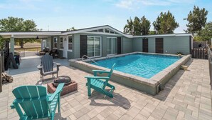 Un área de piscina moderna al aire libre con un porche cubierto, dos sillas Adirondack y una parrilla.