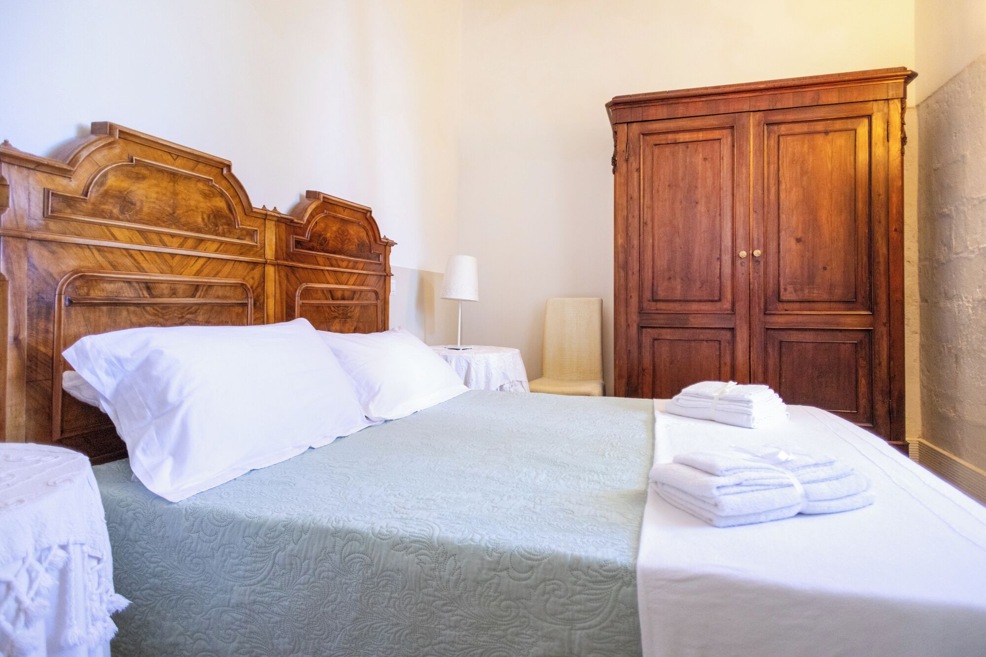 A neatly made bed with white linens, a wooden headboard, and a bedside table with a lamp. There is a wooden wardrobe and a stack of folded towels on the bed.