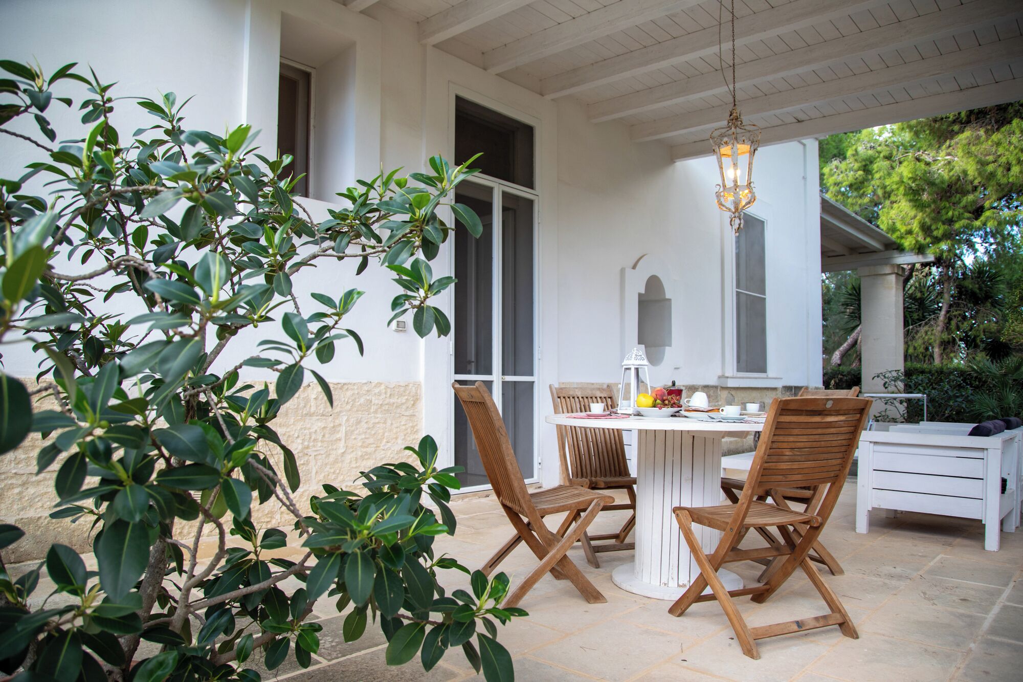 A covered patio with a table and four wooden chairs, surrounded by greenery.