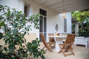 Een overdekt terras met een tafel en vier houten stoelen, omgeven door groen.