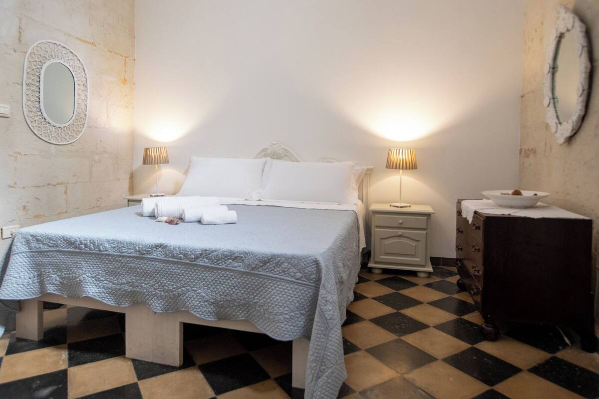 Egy gondosan megterített ágy fehér ágyneművel és kék takaróval, két éjjeli lámpa, egy kis éjjeliszekrény egy tálcal, és kockás mintájú padló.