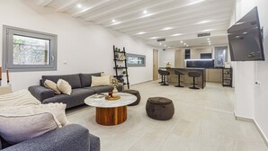 Uma sala de estar moderna com um sofá cinzento, uma mesa de centro redonda e uma área de cozinha ao fundo.