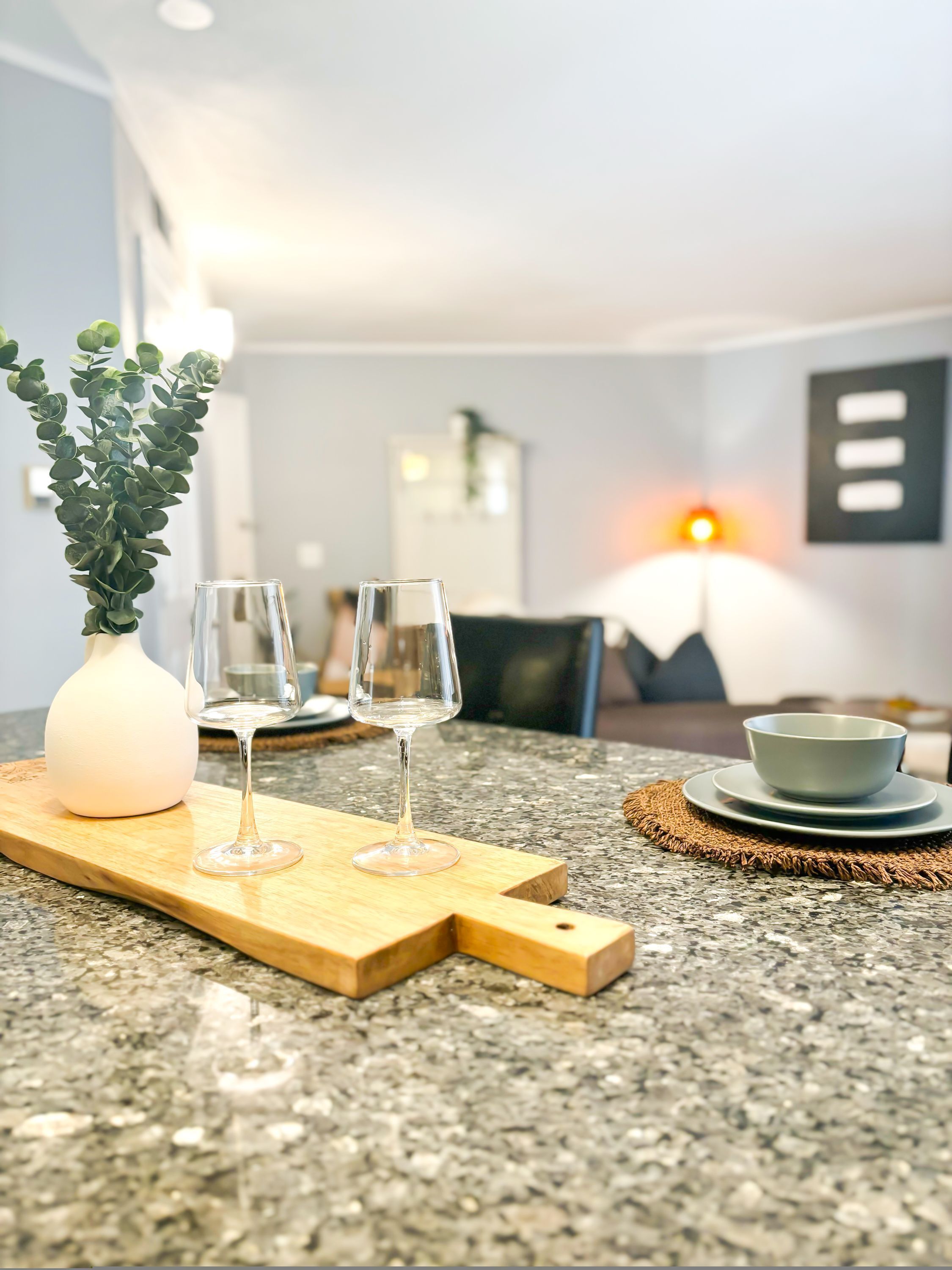 A kitchen counter with a vase of green plants, two wine glasses, and a set of plates and bowls.