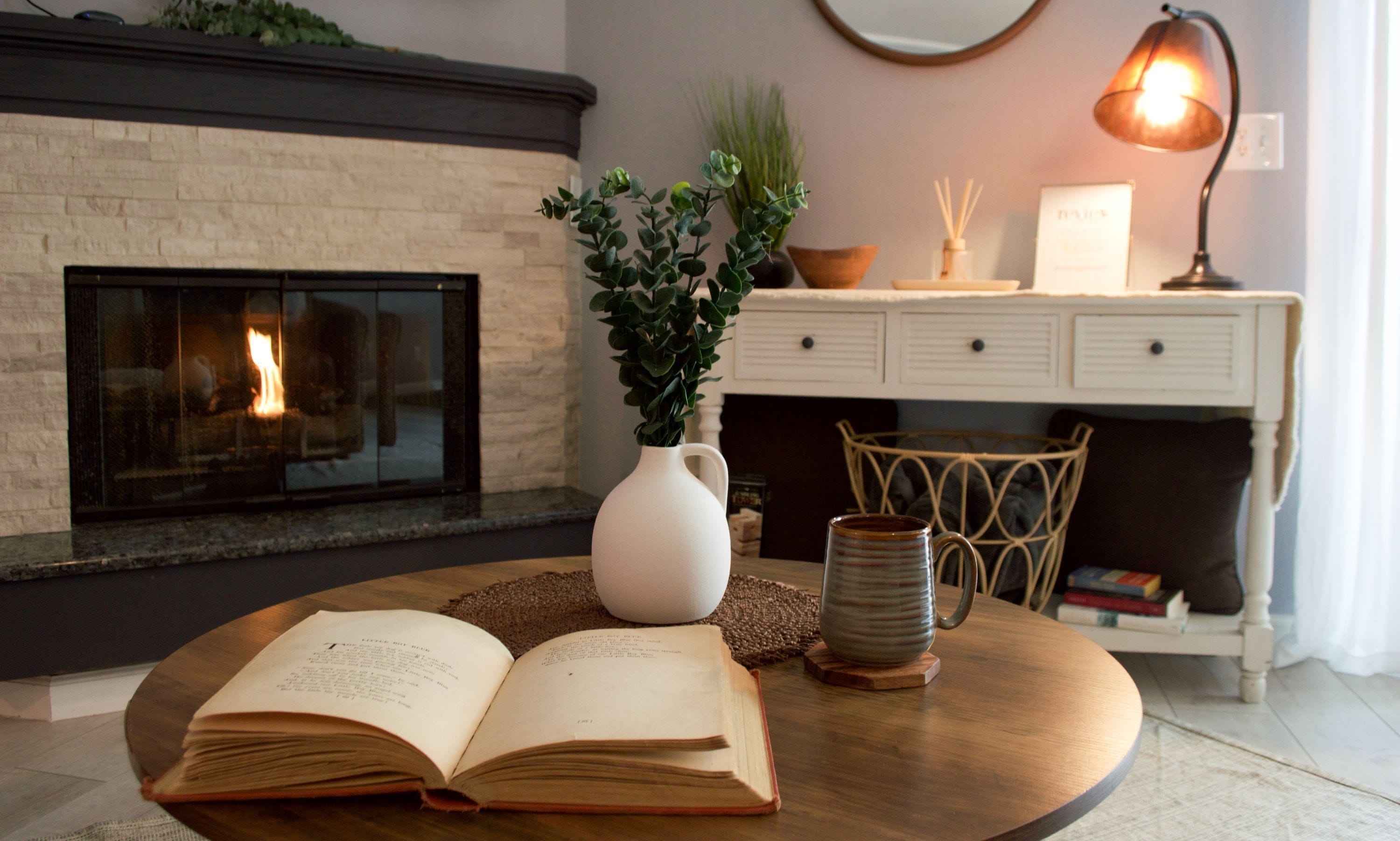 A cozy living room with a fireplace, a round wooden coffee table, an open book, a white vase with green plants, and a wicker chair.