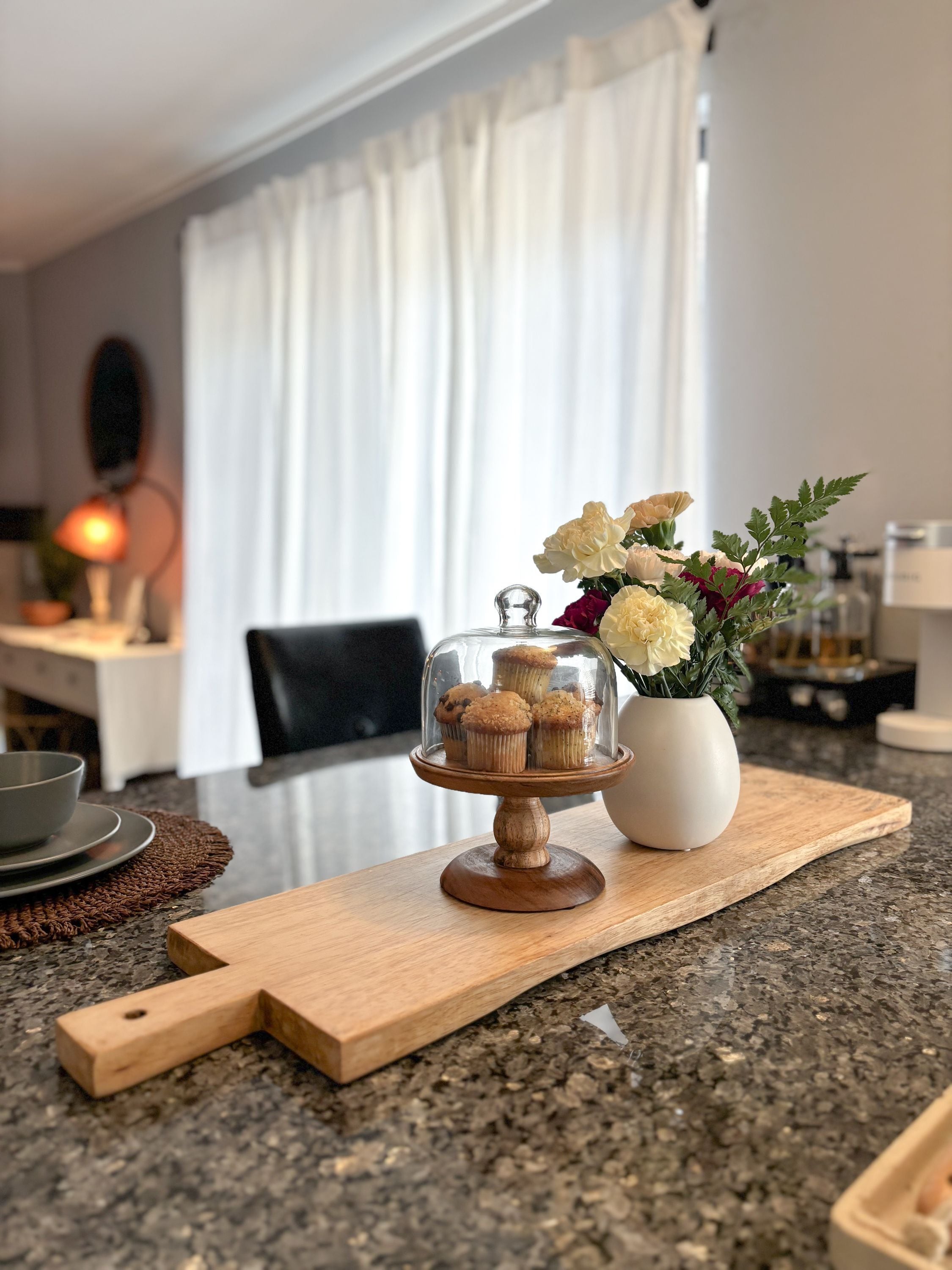 Un comptoir de cuisine avec une planche à découper en bois, un présentoir en verre contenant des muffins, un vase avec des fleurs et un chandelier.