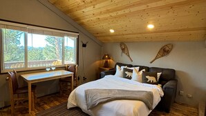 Ein Schlafzimmer mit Holzbalkendecke, einem Bett mit weißer Bettwäsche, einem Schreibtisch mit Stuhl und einem Fenster mit Blick auf Bäume.