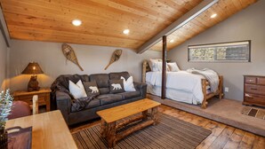 Ein gemütliches Zimmer mit Holzbalkendecke, einer Couch mit Tierkissen, einem Bett mit weißen Leinen und einem Nachttisch aus Holz mit einer Lampe.