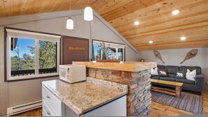 Eine moderne Küche mit Granitarbeitsplatten, einer Steinrückwand und einer eingebauten Mikrowelle. Eine Steinwand, eine Holzbalkendecke und ein Fenster mit Blick auf Bäume sind zu sehen. Im Wohnbereich sind ein graues Sofa und ein Couchtisch aus Holz erkennbar.