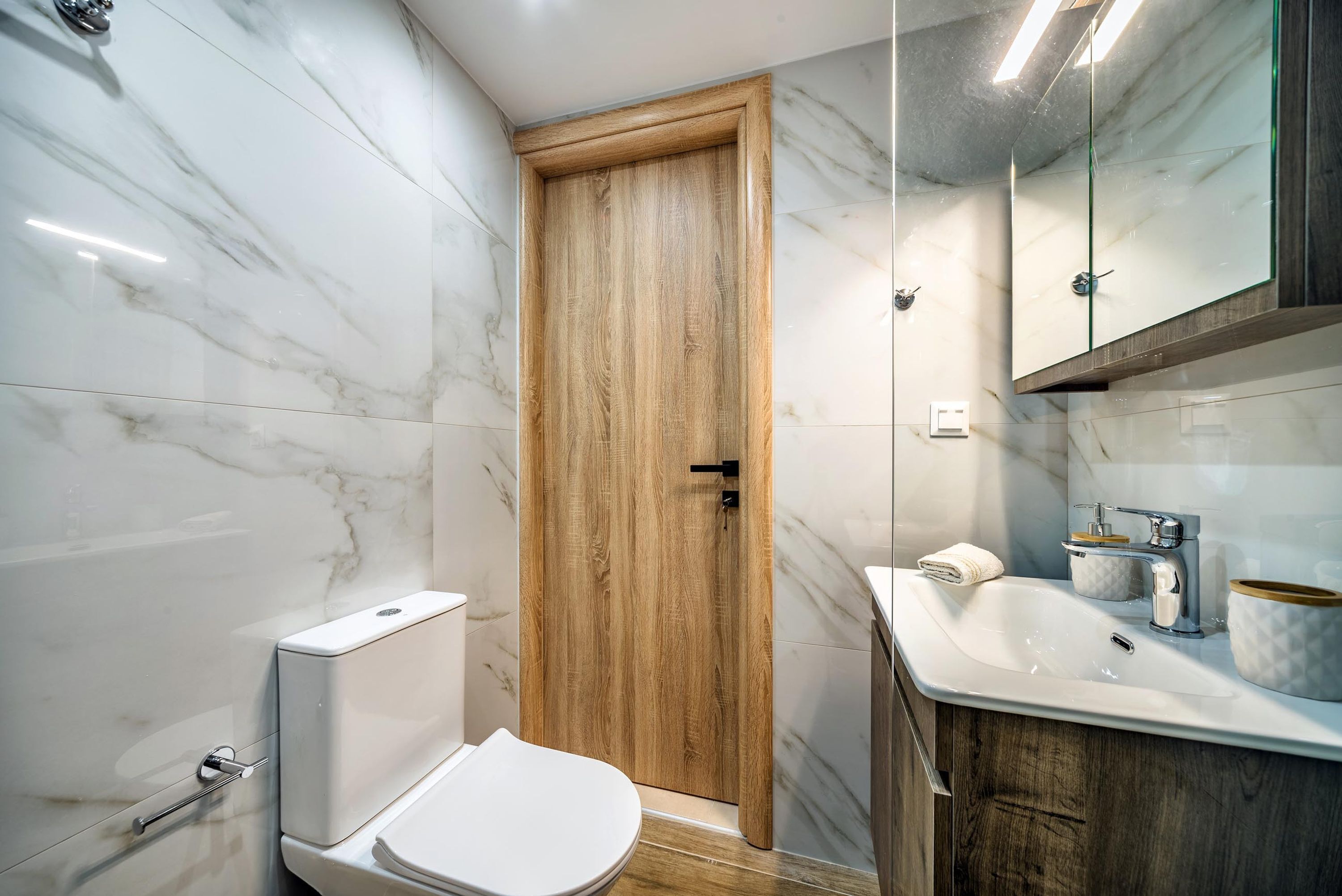 Un baño con puerta de madera, inodoro blanco, lavamanos con espejo y paredes de mármol.