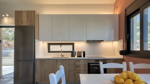 A modern kitchen with a stainless steel refrigerator, wooden cabinets, a white countertop, and a dining table with a plate of lemons. - Lemonia Suites in Ikaria (Ikaria)