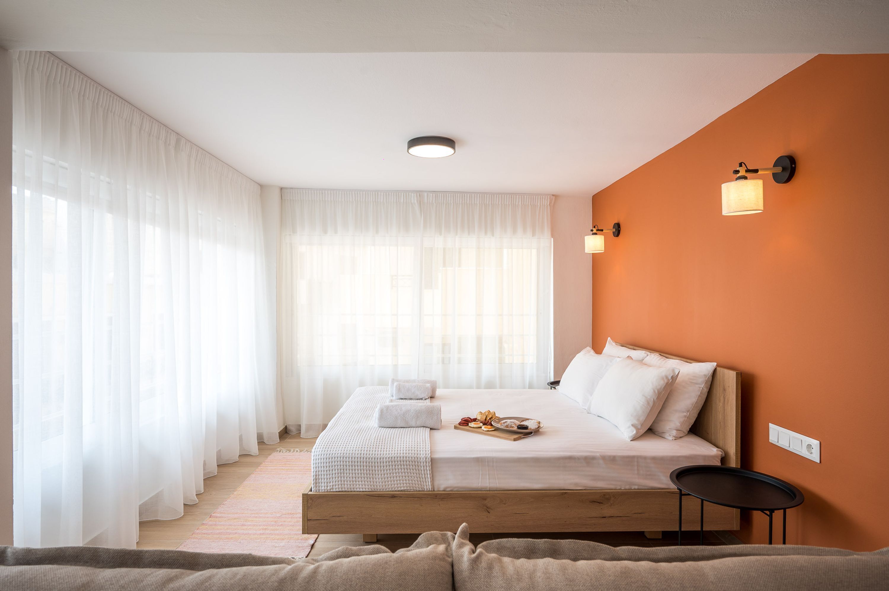 Un dormitorio con cama de madera, ropa de cama blanca, una mesita de noche y una ventana con cortinas de gasa.