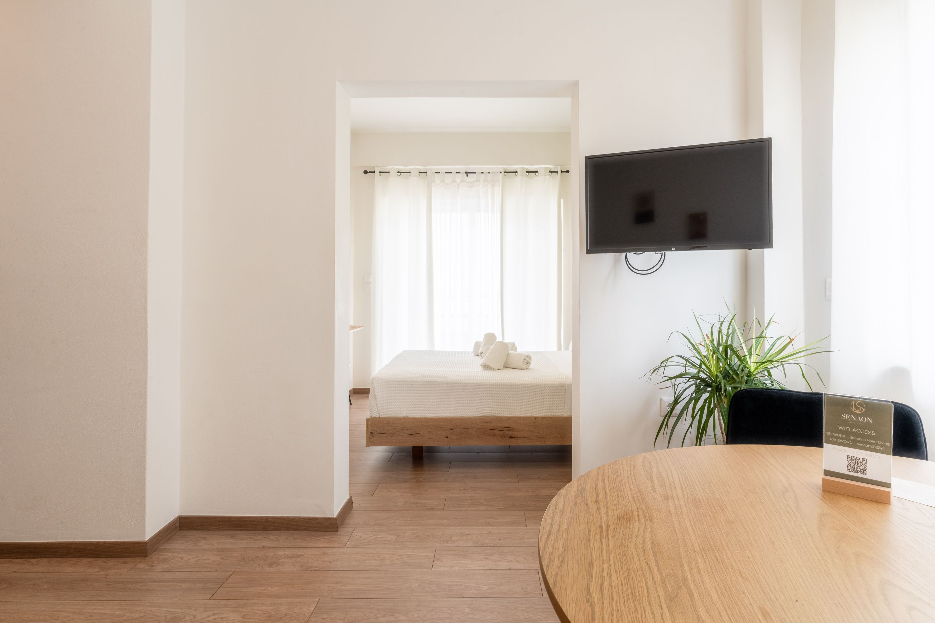 A hotel room with a bed, a TV mounted on the wall, and a wooden table with a sign.