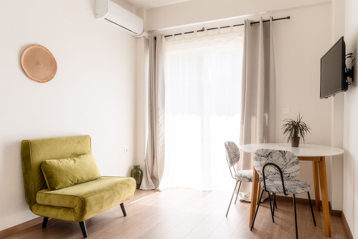 Ein modernes Wohnzimmer mit einem grünen Sofa, einem Esstisch mit Stühlen, einem an der Wand befestigten Fernseher und einem großen Fenster mit Vorhängen.