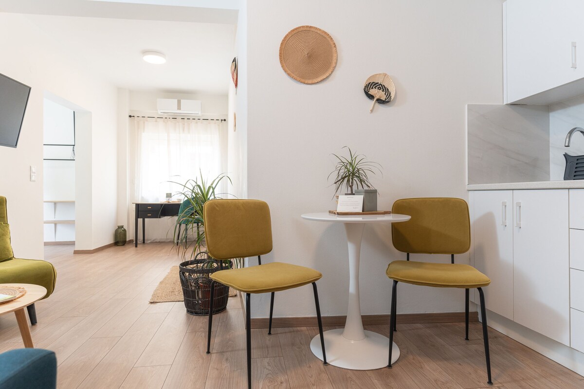 A modern living room with a round white table, two mustard-colored chairs, a green sofa, and a kitchen area with white cabinets and a marble countertop.
