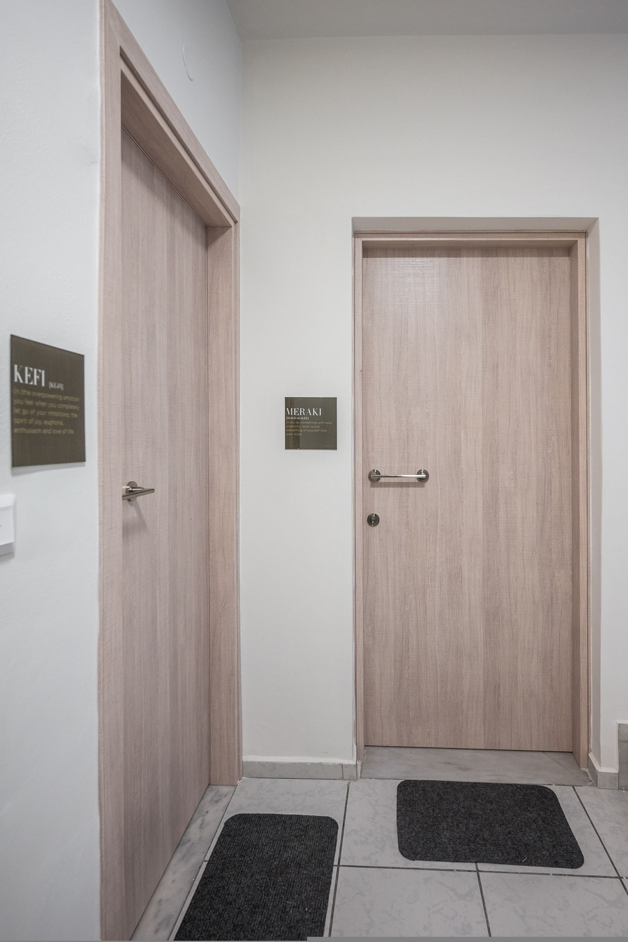 A hallway with two wooden doors, each with a metal handle and a nameplate. There are black doormats on the floor.