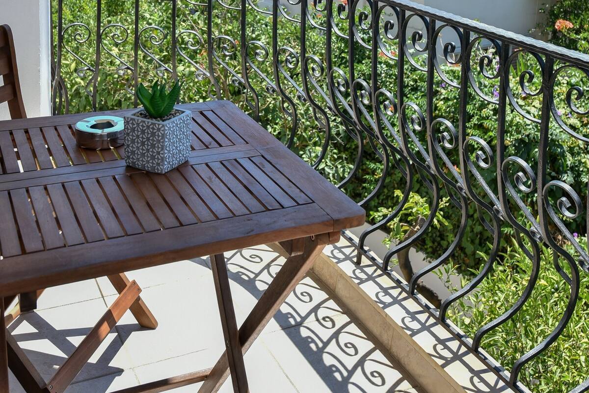 A balcony with a wooden table and chairs, a potted plant, and a view of a hillside with buildings.