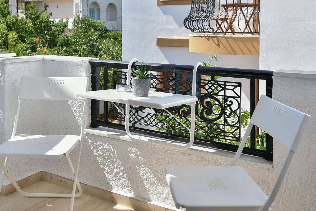 A balcony with white chairs and a small table, overlooking a building with a balcony and a window.