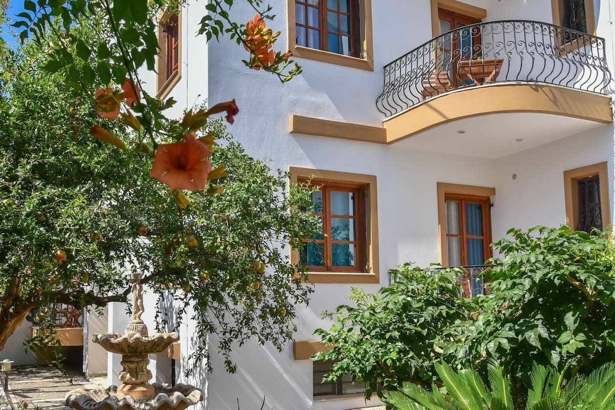 A white building with a balcony, a stone fountain, and lush greenery.